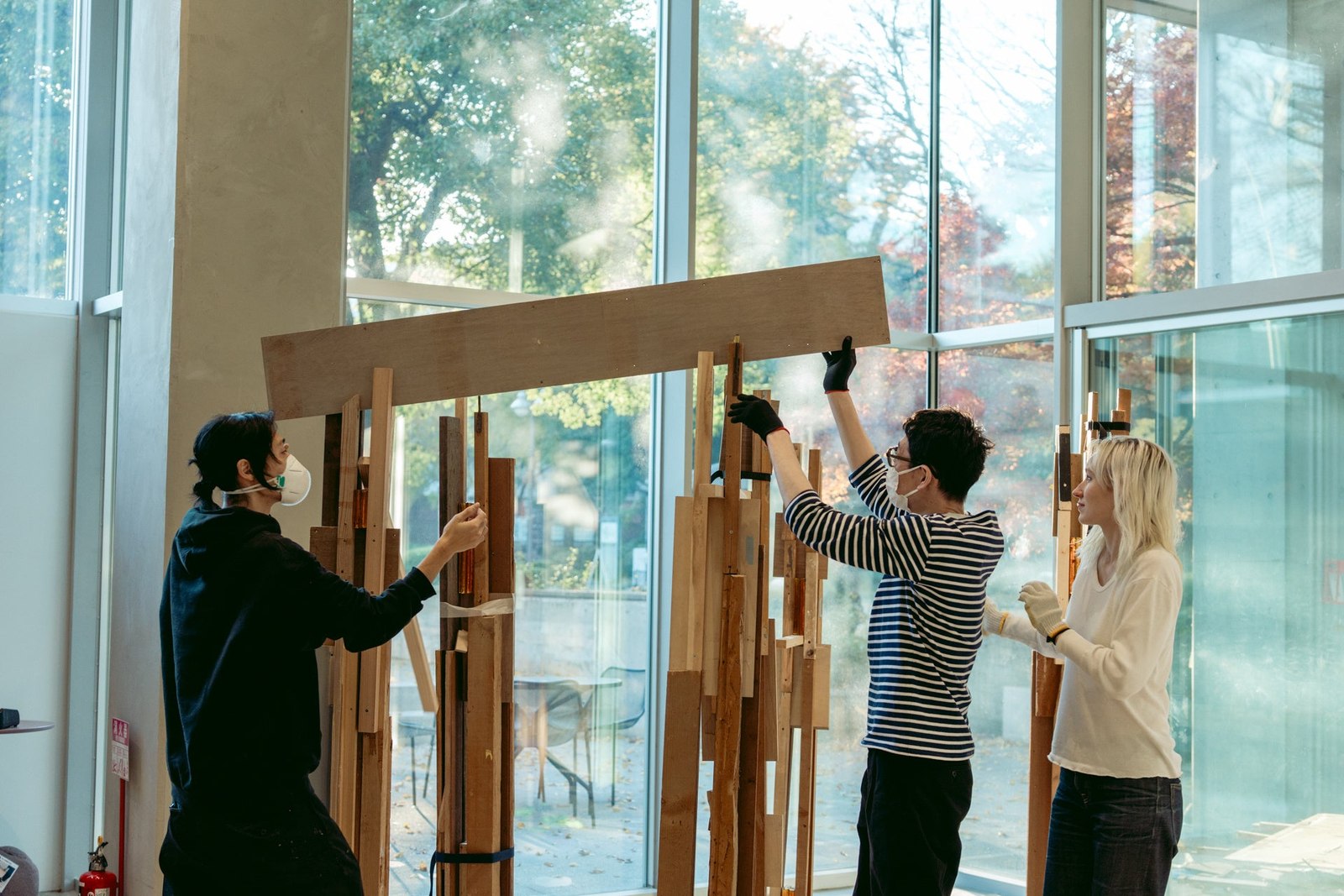 大きな窓のある制作室で、3人が木材を使った作品を組み立てている。
